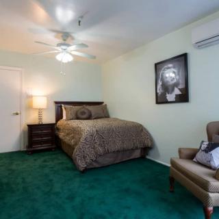 A cozy bedroom featuring a bed, armchair, and artwork on the wall with green carpet flooring.