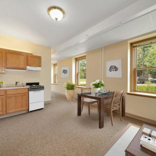 A cozy apartment interior featuring a small kitchen and dining area, with ample natural light from large windows.