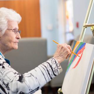 An elderly woman painting on a canvas at an easel.