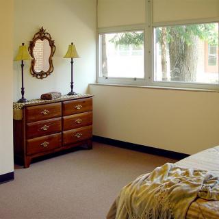 A cozy bedroom with a wooden dresser, decorative mirror, and lamps by the window.