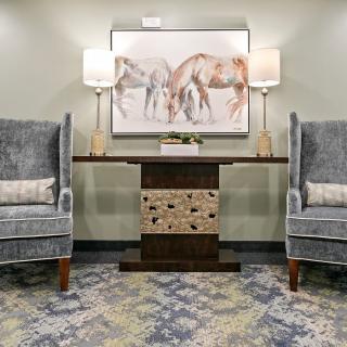 A cozy lobby area featuring two grey armchairs, a console table with drawers, decorative lamps, and a horse-themed painting on the wall.