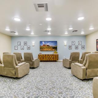 A cozy theater room with beige recliner chairs facing a large television on a patterned carpet floor, surrounded by soft lighting.