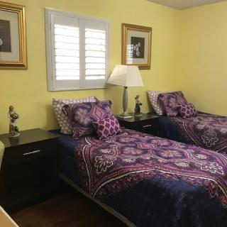 A cozy bedroom with twin beds featuring purple patterned bedding, a central lamp on the nightstand, and framed artwork on the yellow walls.