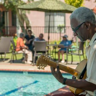 A man plays guitar by the poolside while others relax and enjoy the sunny day.