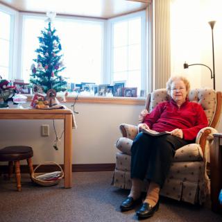 An elderly woman sits in a cozy living room with holiday decorations and a small Christmas tree.