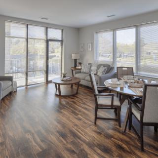 A modern living room with hardwood floors and ample natural light, featuring a cozy seating area and a dining space by large windows.