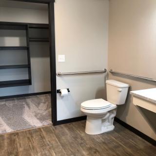Modern accessible bathroom with grab bars, a toilet, sink, and walk-in closet.