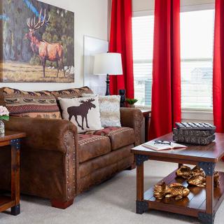 A cozy living room with a brown sofa, featuring decorative pillows and an elk-themed artwork, complemented by red curtains and a wooden coffee table.