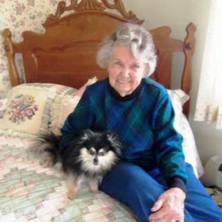 An elderly woman sitting on a bed with her small dog, both looking at the camera and smiling.