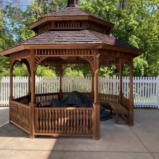 A wooden gazebo surrounded by a white picket fence and greenery.