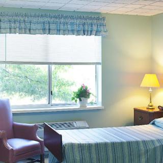 A clean and simple hospital room with a bed next to a window, a lamp on a small table, and a comfortable chair beside the bed.