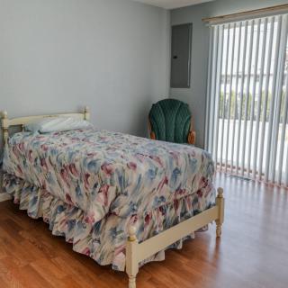 A simple bedroom with a single bed covered in floral bedding, a green chair, and large window blinds letting in daylight.