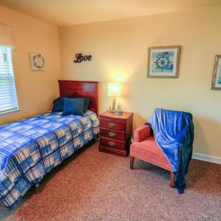 A cozy bedroom with a single bed and an armchair beside a small bedside table, illuminated by a lamp.
