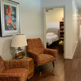 A cozy living space with two armchairs, artwork, and a lamp, leading to a hallway with a bedroom in view.