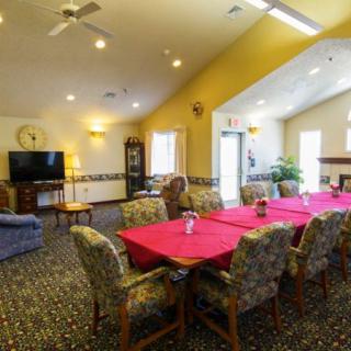 A spacious and well-lit living and dining area featuring a long table with red tablecloths, comfortable chairs, a television, indoor plants, and ample natural light.