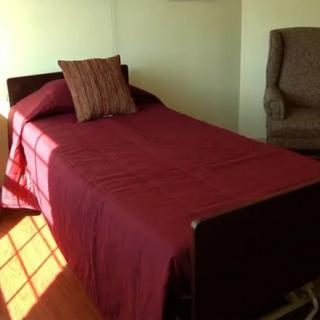 A single bed with a burgundy cover and a pillow, next to an armchair in a well-lit room.