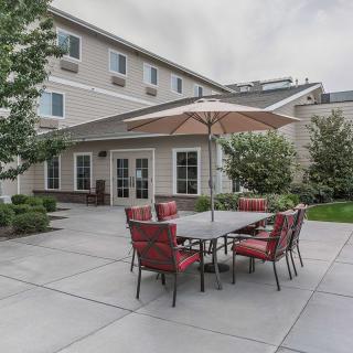 A patio with red cushioned chairs and a table under an umbrella, next to a multi-story building and surrounded by greenery.