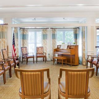 A brightly lit meeting room with arranged chairs and an organ, featuring an American flag by the window.