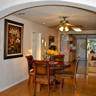 A cozy dining area featuring a round wooden table with chairs, decorated with flowers under a ceiling fan. The wall displays artwork, and the space opens up to a glimpse of the kitchen.