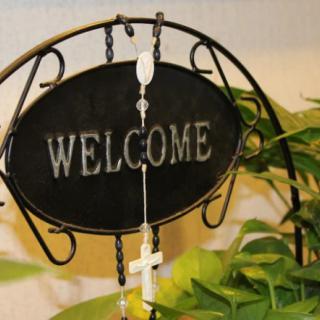 A welcome sign adorned with a rosary and cross, surrounded by indoor plants.