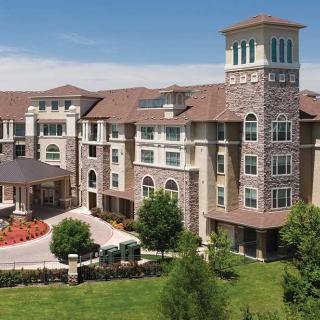 A luxurious multi-story residential building with a tower, surrounded by manicured green lawns and trees under a clear blue sky.