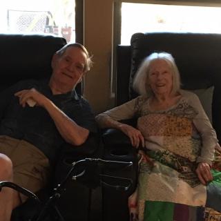 Two elderly individuals sitting together indoors, sharing a moment.