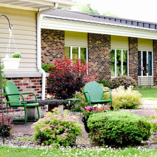 A cozy garden patio with green chairs nestled among lush shrubs and plants in front of a brick house.