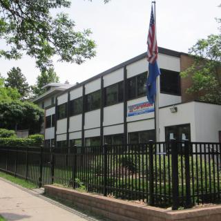 A two-story school building with a flagpole in front, surrounded by a fence and trees.