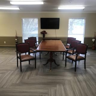 A spacious conference room featuring a large table surrounded by chairs, with a television mounted on the wall and natural light coming through windows.