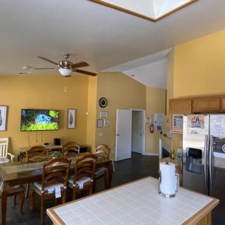 A spacious kitchen and dining area with yellow walls, featuring a ceiling fan, television, dining table with chairs, and stainless steel refrigerator.