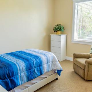 A simple bedroom featuring a single bed with blue bedding, an armchair, and a dresser near a window with natural light.