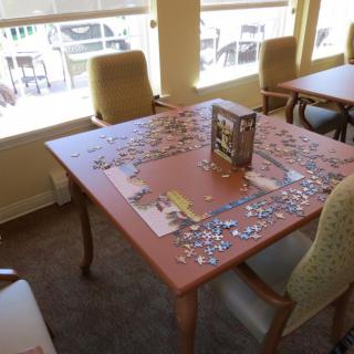 An indoor room featuring a table with a partially completed puzzle, surrounded by chairs and illuminated by natural light from the windows.