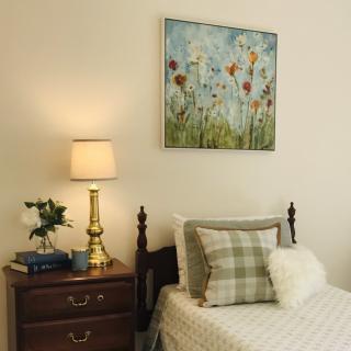 A cozy bedroom with a wooden nightstand, elegant lamp, and a floral painting above the bed.