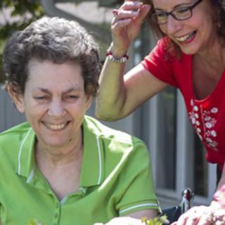 A smiling older adult in a green shirt sits outdoors in a wheelchair with a caregiver in a red shirt nearby.