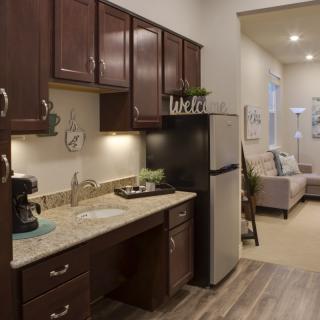 A modern apartment kitchen with wooden cabinets and a small living area in the background.