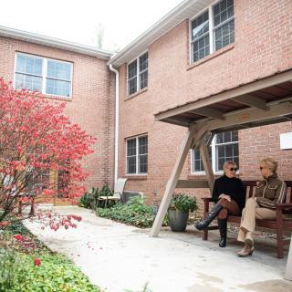 Two people sitting on a bench in an outdoor garden area beside a brick building.