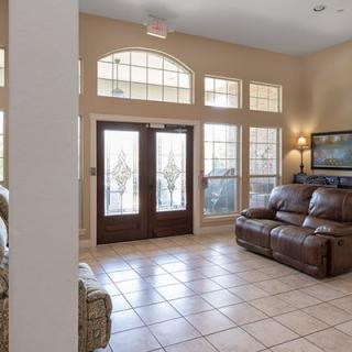 A living room with a tile floor, featuring a sofa, large windows, double doors, and wall artwork.