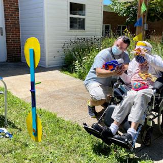A caregiver assists an elderly person in a wheelchair during an outdoor activity, surrounded by vibrant decorations and greenery.