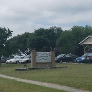 A view of a community area featuring a sign, several parked cars, and landscaped surroundings under an overcast sky.