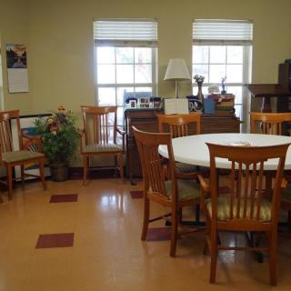A community meeting room with a round table and wooden chairs, surrounded by windows and various furnishings.