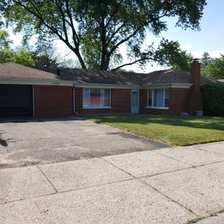 A single-story brick house with a driveway and garage, surrounded by greenery in a suburban neighborhood.