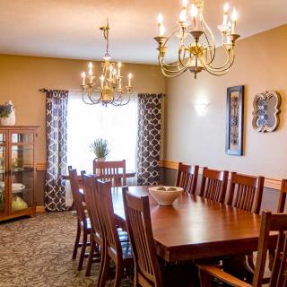 A cozy dining room featuring a wooden table and chairs, illuminated by elegant chandeliers, with decorative elements and natural light filtering through patterned curtains.