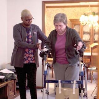 A caregiver assists an elderly woman using a walker in a home setting.