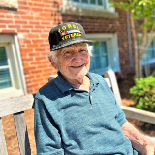 An elderly man wearing a veteran hat sits on a bench, smiling on a sunny day.