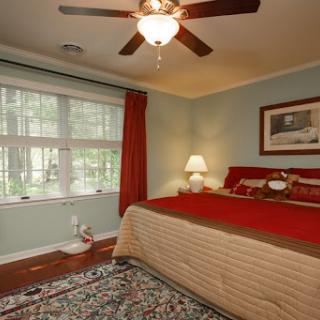 A cozy bedroom featuring red bedding and a ceiling fan, illuminated by natural light from the window.