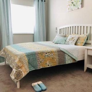 A cozy bedroom featuring a neatly made bed with a colorful quilt, complemented by matching pillows and light blue curtains. Slippers are placed beside the bed.