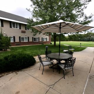 A serene outdoor patio with a table and chairs under an umbrella, adjacent to a building surrounded by greenery.