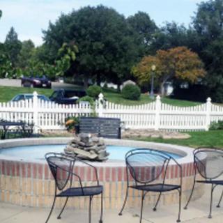 A tranquil outdoor area featuring a fountain surrounded by metal chairs, set against a backdrop of a white picket fence and lush trees.
