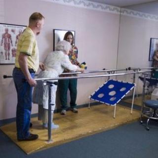 An elderly person receives assistance during a physical therapy session using parallel bars.