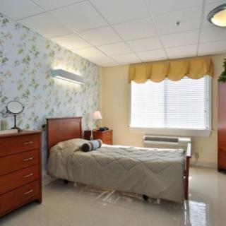 A well-lit room with a single bed and wooden furniture, possibly in a nursing home or hospital setting.
